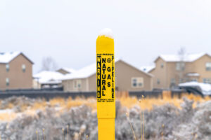 Pole marker with warning sign for natural gas pipeline buried underground. Snowy homes and white sky can be seen in the blurry background landscape.