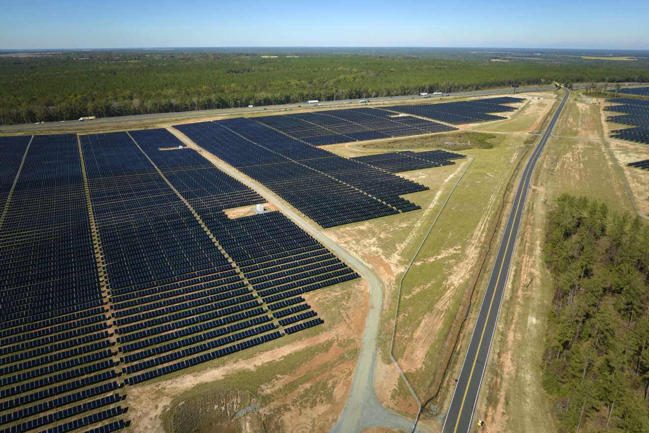 Aerial view of large power plant with rows of solar panels by a highway.